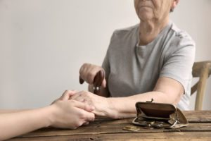 Money coins falling out onto table from open wallet purse as young woman holds hand of elderly woman sitting in chair with walking cane in other hand